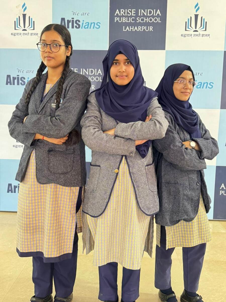 Three confident senior students posing in front of the 'We are Arisians' branded backdrop